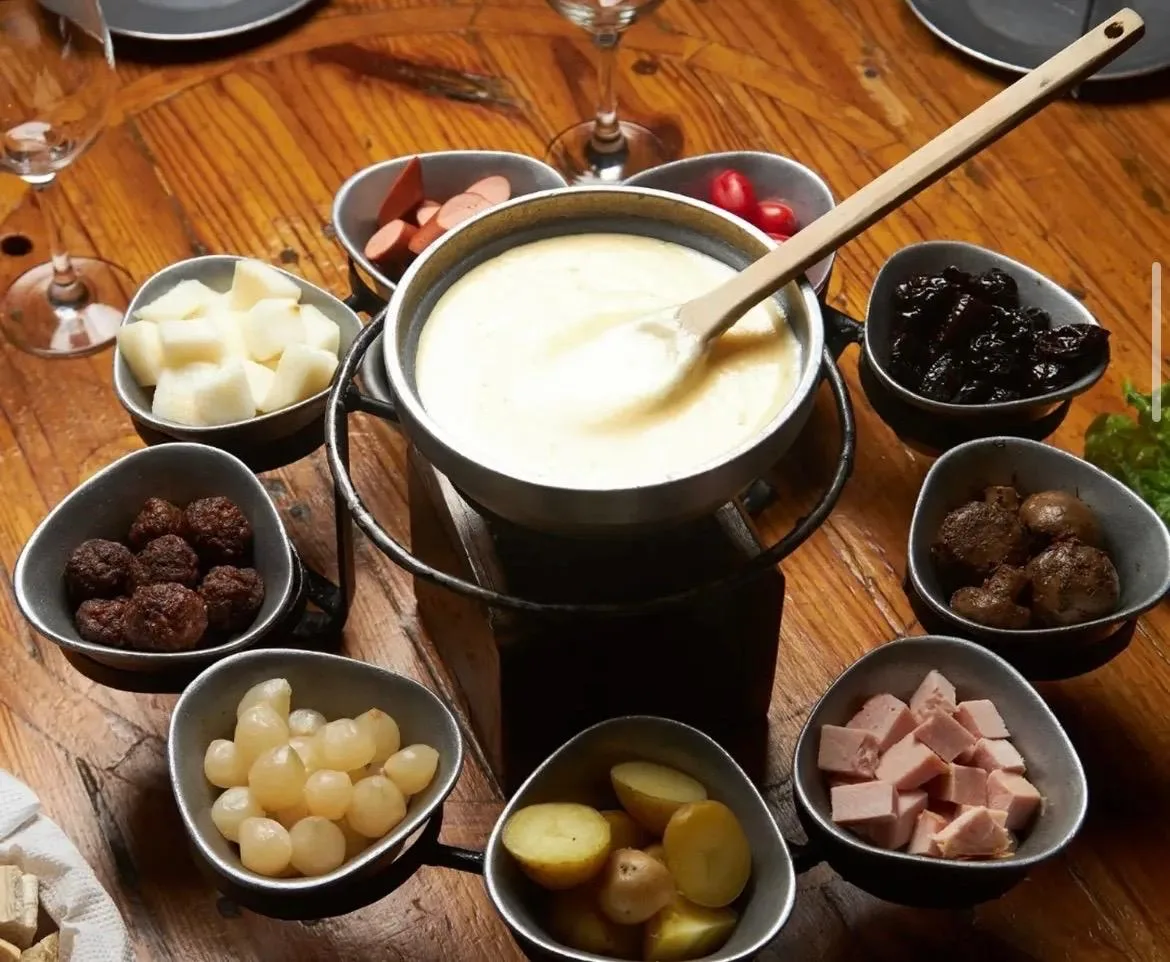 Fondue de queso al centro, rodeada de cazuelitas con papas, pan, fiambres y guarniciones para mojar.