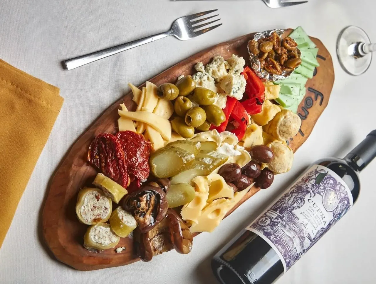 Tabla de fiambres y quesos con aceitunas, morrones y vegetales asados sobre tabla de madera.